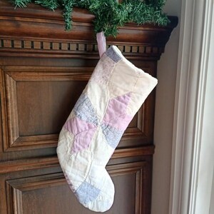 May include: A quilted Christmas stocking featuring a patchwork design of soft pastel colours including light pink, cream, and pale blue. The fabric pieces are stitched together in a star-like pattern with a light pink fabric loop at the top for hanging.