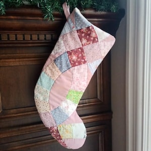 Peut inclure: Une chaussette de Noël faite à la main avec un motif patchwork. La chaussette présente divers tissus dans des tons de rose, de rouge, de bleu et de vert, disposés en motif matelassé. Elle est suspendue sur un fond en bois foncé.