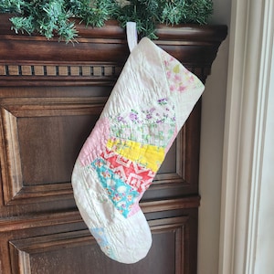 May include: A quilted Christmas stocking featuring a patchwork design of various floral and geometric fabric scraps in shades of white, pink, yellow, red, and blue, finished with a white fabric hanging loop at the top edge.