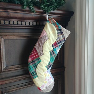 May include: A patchwork Christmas stocking with a yellow, green, red and brown plaid design. The stocking is hanging on a wooden mantelpiece.