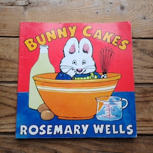 May include: A children's book titled "Bunny Cakes" by Rosemary Wells. The cover features a white bunny in a mixing bowl, a bottle, eggs, and a measuring cup. The background is red and blue.