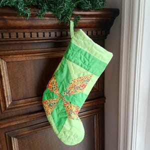 Peut inclure: Une chaussette de Noël matelassée verte avec un motif floral, suspendue à une cheminée en bois. Le motif de la chaussette comprend un motif central en forme d'étoile. Une décoration festive pour les fêtes de fin d'année.