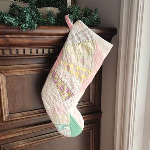 Könnte beinhalten: Ein gesteppter Weihnachtsstrumpf mit einem Patchwork-Design aus verschiedenen pastellfarbenen Stoffen, darunter hellrosa, gelbe und grüne Blumenmuster. Der Strumpf hat eine weiche, gesteppte Textur und eine kleine Schlaufe aus rosa Stoff oben.