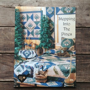 May include: A quilt pattern book titled "Stepping Into The Pines" featuring a quilt with a pine tree design. The book cover shows a bedroom with a bed made with the quilt, a pine tree, and a teddy bear.
