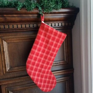 Peut inclure: Chaussette de Noël à carreaux rouges et blancs suspendue à une cheminée en bois foncé. La chaussette est en tissu tissé avec un motif de vacances classique. Une guirlande verte est drapée au-dessus du manteau.