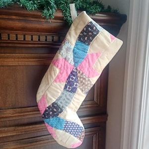 May include: A quilted Christmas stocking featuring a patchwork design with a central star motif. The fabric pieces include solid beige, bright pink, and various floral and paisley patterns in shades of blue, brown, and black with a hanging loop.