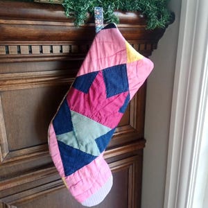 May include: A patchwork Christmas stocking in shades of pink, blue, yellow, and green. The design features a starburst pattern created from various fabric pieces. A floral fabric loop is attached for hanging the stocking.