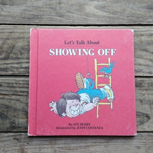 May include: A red book titled "Let's Talk About Showing Off" with an illustration of a boy hanging upside down from a ladder. The book is by Joy Berry and illustrated by John Costanza.