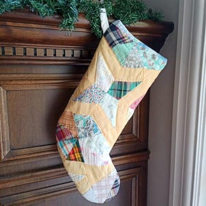 May include: A quilted Christmas stocking featuring a patchwork design of various fabric patterns including plaid, floral, and solid yellow sections. The stocking is constructed with a star-shaped motif and includes a white fabric loop for hanging.