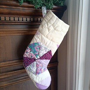 May include: A patchwork Christmas stocking in cream, purple, and green. The stocking has a quilted design with a floral pattern and a purple toe. It is hanging against a wooden background, ready for festive treats.