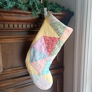 May include: A quilted Christmas stocking featuring a patchwork design of various pastel-coloured fabrics including yellow, pink, light blue, and green with floral and geometric patterns. The stocking has a white fabric loop at the top for hanging.