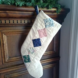Könnte beinhalten: Ein gesteppter, cremefarbener Weihnachtsstrumpf mit einer vertikalen Reihe aus fünf rautenförmigen Stoffaufnähern. Die Aufnäher zeigen florale Muster in Blau, Grün und Weiß, ein einfarbiges Altrosa-Quadrat sowie ein dunkelblaues Quadrat mit Muster.