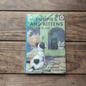 May include: A vintage children's book titled "Puppies and Kittens" by M.E. Gagg. The cover features a black and white puppy looking at a black cat and a white kitten through a doghouse door. The book is part of the "A Ladybird Learning to Read Book" series.