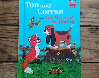 May include: A vintage children's book titled "Tod and Copper" from "The Fox and the Hound" with a teal cover. The cover features illustrations of a red fox, a brown dog, a butterfly, and a turtle. The book is set against a wooden background.