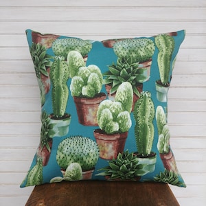 May include: Teal blue throw pillow with a repeating pattern of various cacti and succulents in terracotta pots. The pillow is made of fabric and has a soft, comfortable feel.