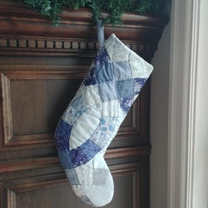 May include: A blue and white patchwork Christmas stocking with a quilted design. The stocking is hanging on a wooden door.