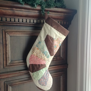 Peut inclure: Une chaussette de Noël en patchwork avec des tissus marron, blanc et fleuris. La chaussette est accrochée à une surface en bois foncé.