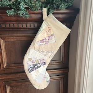 Könnte beinhalten: Ein gesteppter Weihnachtsstrumpf mit einem fächerförmigen Patchwork-Design auf der Vorderseite. Die Stoffbahnen zeigen verschiedene florale und abstrakte Muster in Violett-, Creme-, Beige- und Weißtönen, abgerundet durch ein Bündchen aus beigem Stoff.