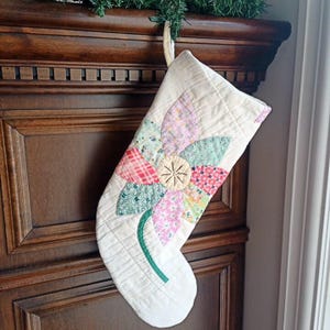 Peut inclure: Chaussette de Noël crème avec une grande fleur colorée appliquée. La fleur présente des pétales en tissu à motifs roses, verts et rouges et un centre jaune. La chaussette est matelassée et possède une tige verte. Une boucle en corde est attachée pour l'accrocher.