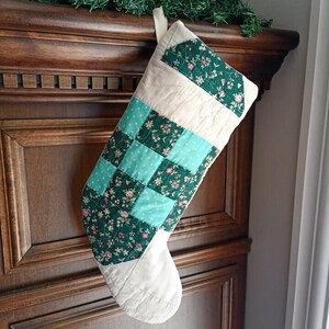 Könnte beinhalten: Ein gesteppter Weihnachtsstrumpf mit einem Patchwork-Design aus abwechselnden dunkelgrünen Blumenstoffquadraten und helltürkisfarbenen gepunkteten Quadraten, akzentuiert durch cremeweiße Stoffabschnitte am oberen Bündchen und an der Fußspitze.