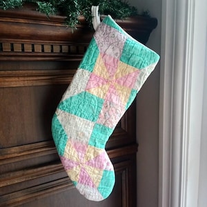 May include: A quilted Christmas stocking featuring a patchwork design of geometric star patterns. The fabric squares are coloured in soft shades of mint green, pale pink, cream, and yellow, with a white fabric loop at the top for hanging.