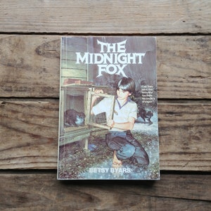 May include: A vintage paperback book cover featuring an illustration of a young boy holding a cage with a fox inside. The book title is "The Midnight Fox" and the author is Betsy Byars.