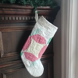 May include: A quilted Christmas stocking with a patchwork design in white, pink, green, and cream colours. The stocking is hanging from a dark wooden mantelpiece, with a green garland above.