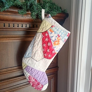 May include: A quilted Christmas stocking constructed from various fabric patches in shades of red, yellow, light blue, and lavender. The stocking features a fan-shaped patchwork design with white floral embroidery on the red sections and a fabric loop.
