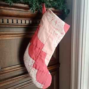 May include: A patchwork Christmas stocking in pink and red hues, featuring a floral design on a cream backdrop. It's suspended from a wooden surface, with a green garland above.