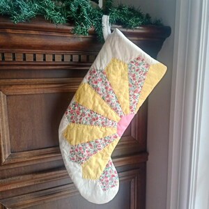 May include: A quilted Christmas stocking featuring a fan-shaped pattern with alternating segments of solid yellow fabric and a floral print fabric with pink and green flowers on a white background, finished with a white fabric cuff and hanging loop.
