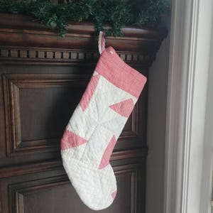 May include: A white quilted Christmas stocking with red and white gingham trim. The stocking has a star-shaped design and is hanging on a wooden surface.