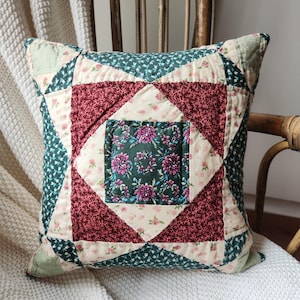 May include: A square quilted throw pillow featuring a patchwork design with a central dark green square adorned with pink floral patterns, surrounded by alternating cream and deep maroon fabric sections with intricate stitched detailing.