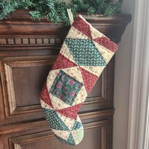 May include: A quilted Christmas stocking featuring a patchwork design of cream, dark red, and forest green fabric panels. The fabric displays floral and geometric patterns throughout, with a fabric loop at the top for hanging purposes.