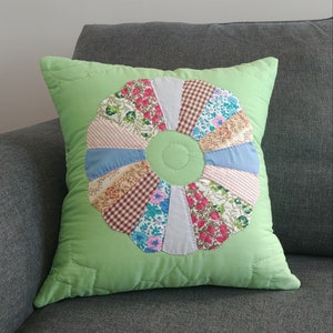 May include: A green cushion with a colourful patchwork flower design in the centre. The flower is made of various fabrics in shades of pink, blue, brown, and white.
