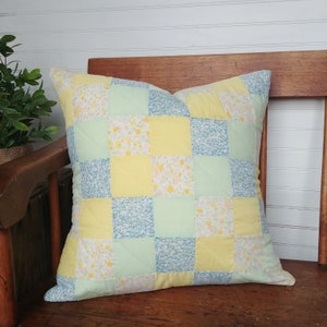 May include: A patchwork cushion with squares of yellow, light green, and blue floral fabric. The cushion is sitting on a wooden bench.