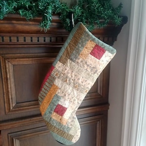 May include: A quilted Christmas stocking featuring a log cabin patchwork design. The fabric panels consist of various muted earth tones including tan, olive green, and mustard yellow, accented by small squares of solid deep red fabric throughout.