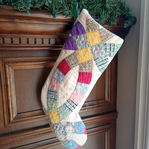 May include: A quilted Christmas stocking with a patchwork design. The stocking features squares and curved sections in various colors, including purple, red, yellow, and blue, set against a cream background. A green garland is visible above the stocking.