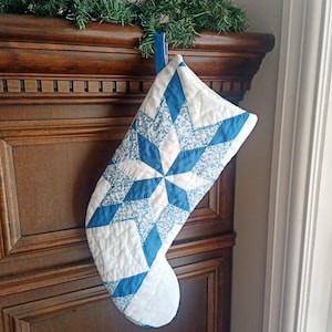 May include: A blue and white quilted Christmas stocking with a starburst pattern. The stocking is made of fabric and has a loop for hanging. The starburst design is made of blue diamonds and white fabric.