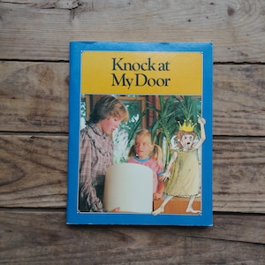 May include: A blue and yellow book cover with the title "Knock at My Door". The cover features an illustration of three children, one wearing a crown, standing in front of a window.