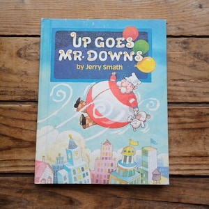 May include: A children's book titled "Up Goes Mr. Downs" by Jerry Smath. The cover features an illustration of a man in a chef's hat floating above a city, held up by balloons. The color palette includes blue, red, yellow, and green.