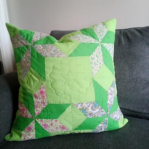 Peut inclure: Un coussin carré fait à la main avec un motif patchwork. Le coussin présente différentes nuances de vert, avec des motifs floraux et géométriques. Le centre présente un motif d'étoile matelassée. Le coussin repose sur un canapé gris.