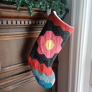 May include: A handmade Christmas stocking with a patchwork design. The stocking features a large flower in the center, with red, black, and blue geometric shapes. The flower has a yellow center. The stocking is hanging from a wooden mantel.