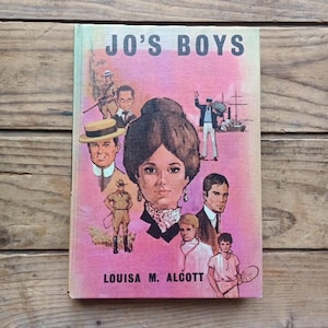 May include: A vintage copy of "Jo's Boys" by Louisa M. Alcott. The book cover is illustrated with several figures, including a woman with a large hairstyle, and the title in bold letters. The background is a pink and peach colour.