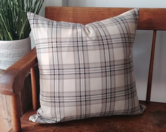 May include: A square decorative pillow with a plaid pattern in shades of beige, gray, and navy blue. The pillow is placed on a wooden bench. A potted plant is visible in the background.