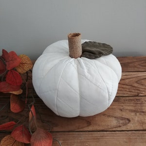 May include: A white quilted pumpkin with a brown hessian stem and a green fabric leaf. The pumpkin is sitting on a wooden surface.