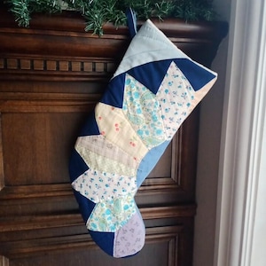 May include: A handmade Christmas stocking with a patchwork design. The stocking features various fabrics in shades of blue, cream, and floral patterns. It has a loop for hanging and is displayed against a dark wooden background.