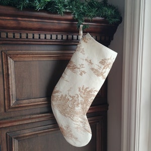 May include: A white Christmas stocking with a brown toile pattern hangs on a wooden mantelpiece. The stocking features a repeating pattern of trees, birds, and houses.