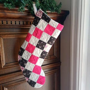 Peut inclure: Une chaussette de Noël matelassée présentant un motif en patchwork composé de carrés alternés en rose vif uni, crème et un tissu marron foncé avec un petit imprimé floral blanc. La chaussette possède une boucle de suspension et une texture matelassée.