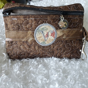 May include: A brown quilted zippered pouch with a silver circle charm featuring the letter "V" and a small silver charm with a paw print. The pouch has a brown and white floral pattern.