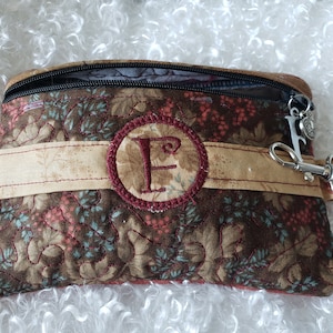 May include: A small brown zippered pouch with a floral pattern and a red embroidered circle with the letter "F" in the center. The pouch has a beige fabric strap and a silver key chain attached.